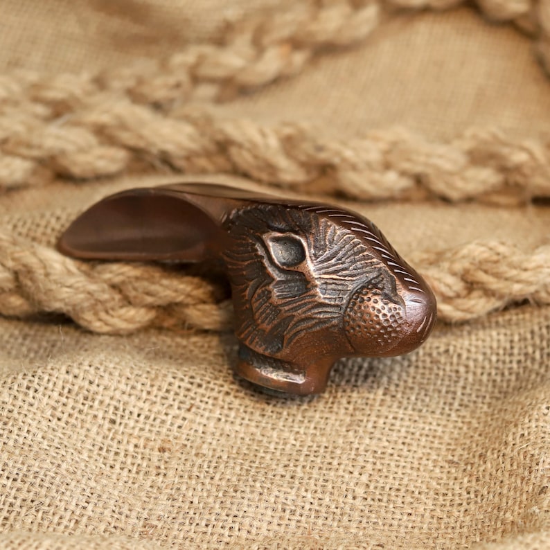 May include: A bronze-colored, decorative door handle shaped like a rabbit's head. The handle features detailed carvings of the rabbit's fur and facial features. The handle is set against a backdrop of burlap and rope.