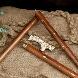 K&ouml;nnte beinhalten: Zerlegte Holzgehstock mit dekorativem goldfarbenem Griff. Die Holzabschnitte des Stocks sind braun mit goldfarbenen Akzenten. Der Griff weist ein verziertes, graviertes Design auf. Der Stock liegt auf einer strukturierten, beigefarbenen Oberfl&auml;che.