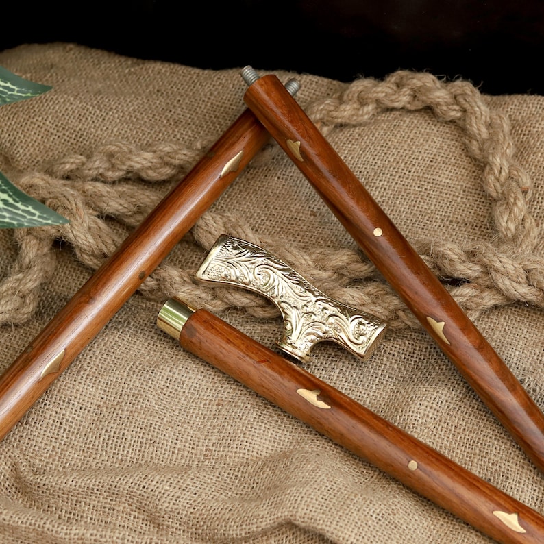 K&ouml;nnte beinhalten: Zerlegte Holzgehstock mit dekorativem goldfarbenem Griff. Die Holzabschnitte des Stocks sind braun mit goldfarbenen Akzenten. Der Griff weist ein verziertes, graviertes Design auf. Der Stock liegt auf einer strukturierten, beigefarbenen Oberfl&auml;che.