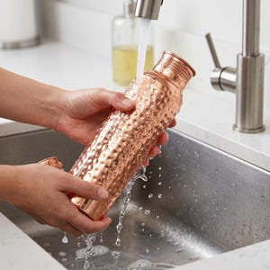May include: A copper water bottle with a hammered texture is held under running water in a stainless steel sink. The bottle is cylindrical with a screw-on cap. The copper has a warm, reddish-brown hue.