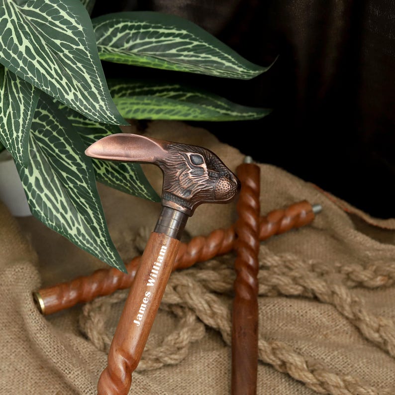 May include: A wooden walking cane with a bronze rabbit head handle. The cane has a twisted design and the name "James William" is engraved on the shaft. Two additional canes are in the background.