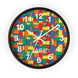 Puede incluir: Un reloj de pared redondo con marco negro. La esfera del reloj presenta un patrón colorido de bloques de construcción de juguete en rojo, amarillo, azul y naranja. Los números blancos marcan las horas y las manecillas son blancas. El nombre "JACK" está impreso debajo.