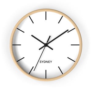 May include: Round wall clock with a light wood frame and a white face. Black hands and hour markers contrast against the white background. The word "SYDNEY" is printed in black at the bottom of the clock face.