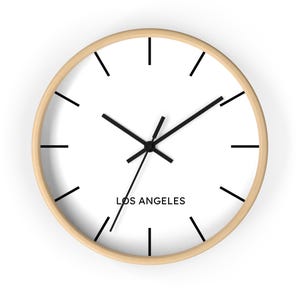 May include: A round wall clock with a light wood frame and a white face. Black hands and hour markers contrast against the white background. The text "LOS ANGELES" is printed in black at the bottom.