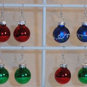 May include: Six pairs of Christmas ornament earrings. Two pairs are red, two are green, and two are blue with white lights painted on them.