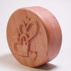 May include: A round wooden box with a carved design of two intertwined hearts. The box is made of a light brown wood and has a smooth finish.
