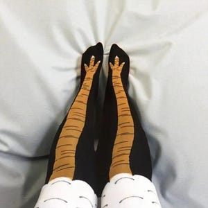 May include: Black novelty socks with a chicken leg design. The socks feature a brown and tan chicken leg graphic on the legs and white chicken feet on the toes. The socks are shown on a light blue surface.