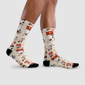 以下が含まれることがあります： ベージュ色の靴下。スヌーピーのキャラクターがフェラーリの旗を持っている模様が繰り返されています。靴下のかかととつま先は黒で、チェッカーフラッグとハートのアクセントがあります。全体的に遊び心のあるカラフルなデザインです。