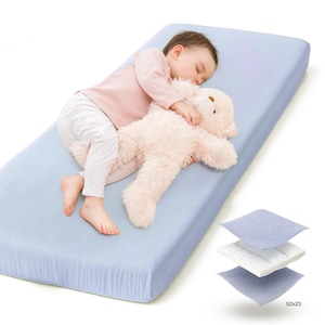 May include: A baby sleeping on a light blue mattress, hugging a plush teddy bear. The mattress is rectangular, with dimensions of 52 x 23 inches. Three fabric samples are displayed, showcasing the mattress's layers.