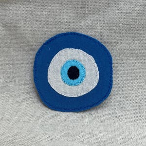Op de afbeelding: Een ronde stoffen patch met een boze oog-ontwerp. De patch bestaat uit een donkerblauwe buitenring, een witte middelste ring, een lichtblauwe binnenste cirkel en een effen zwart midden, samengevoegd met zichtbare blauwe stiksels.