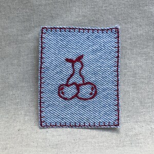 May include: A rectangular light blue denim patch featuring a dark red embroidered cherry design in the center. The edges of the patch are finished with a dark red blanket stitch border, creating a distinct contrast against the blue fabric.