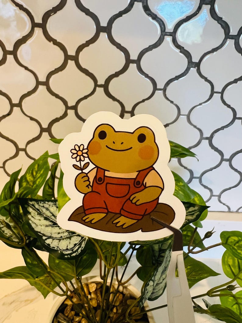 May include: A sticker featuring a cartoon frog wearing orange overalls, holding a daisy. The frog is sitting on a brown leaf. The sticker has a white border and is placed on a plant with green leaves.