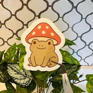 May include: A sticker featuring a cartoon frog wearing a mushroom cap. The frog is light brown with rosy cheeks, and the mushroom is red with white spots. The sticker is held by tweezers in front of a plant with green leaves.