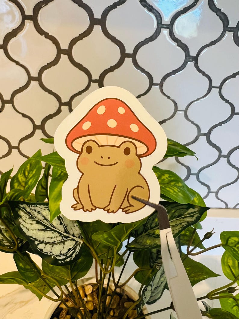 May include: A sticker featuring a cartoon frog wearing a mushroom cap. The frog is light brown with rosy cheeks, and the mushroom is red with white spots. The sticker is held by tweezers in front of a plant with green leaves.