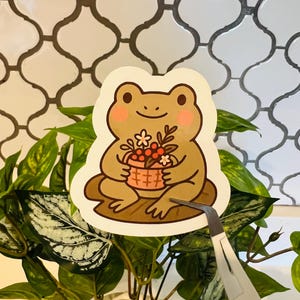 May include: A sticker featuring a cartoon frog holding a basket of flowers. The frog is light brown with pink cheeks and is sitting on a brown lily pad. The sticker is being held by tweezers in front of a plant.