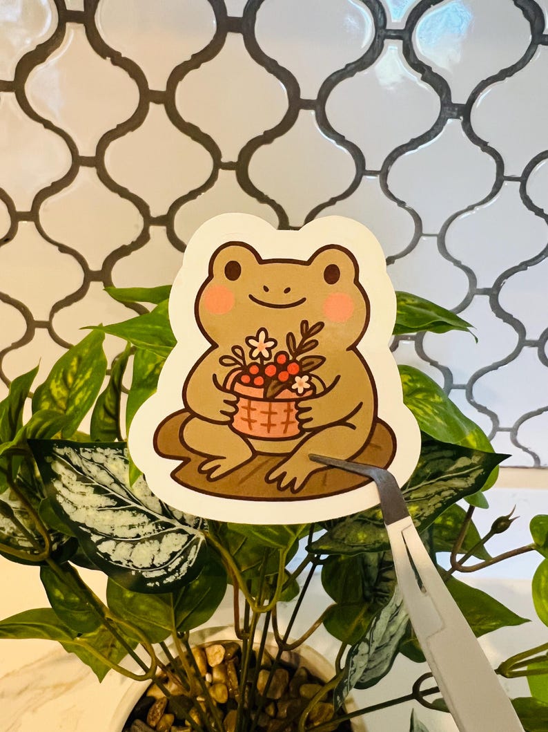 May include: A sticker featuring a cartoon frog holding a basket of flowers. The frog is light brown with pink cheeks and is sitting on a brown lily pad. The sticker is being held by tweezers in front of a plant.