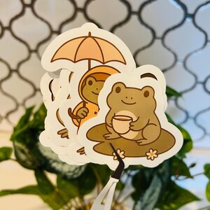 May include: A collection of frog-themed stickers. One sticker features a frog under an umbrella, another shows a frog in a raincoat, and a third depicts a frog holding a cup. The stickers have a white border and are held by tweezers.