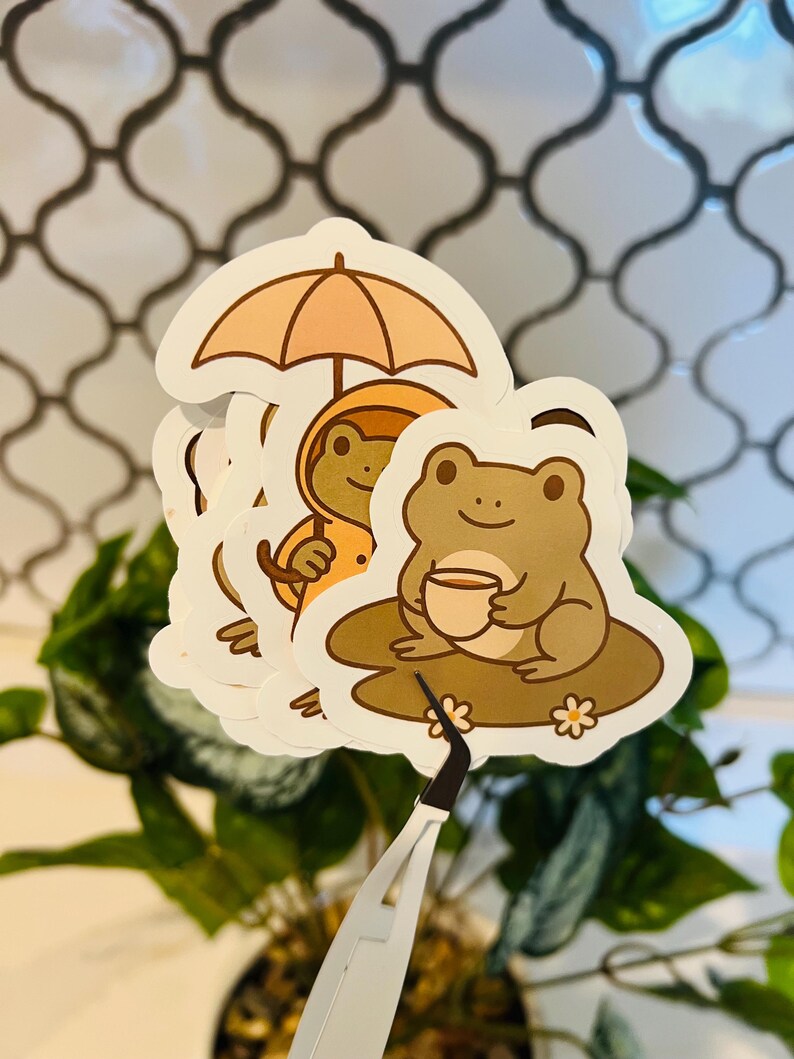 May include: A collection of frog-themed stickers. One sticker features a frog under an umbrella, another shows a frog in a raincoat, and a third depicts a frog holding a cup. The stickers have a white border and are held by tweezers.