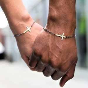 May include: Two gold cross bracelets on dark gray chain. The bracelets are worn on wrists, with the hands clasped together. The cross is a simple, minimalist design. The background is blurred, suggesting an outdoor setting.