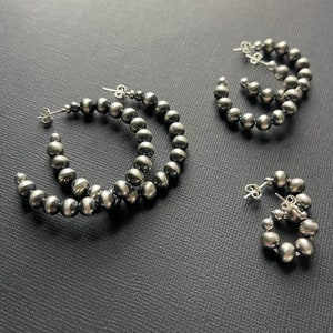 May include: Three pairs of silver hoop earrings with a beaded design. The earrings are made of small, round beads that are connected together to form a hoop shape. The beads are a dark silver color.