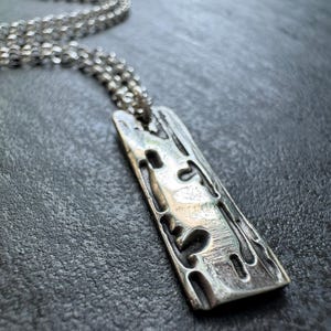 Peut inclure: Un collier pendentif argenté avec un pendentif rectangulaire texturé. Le pendentif présente un design ajouré et sculpté avec des formes organiques. La chaîne est de style délicat et maillé. Le collier repose sur une surface gris foncé.