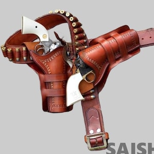 May include: A brown leather double holster gun belt featuring intricate decorative stitching, two silver-toned revolvers with white grips, and multiple brass-cased cartridges held in loops along the belt and attached to the holster assembly.