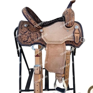 May include: A Western-style leather saddle with intricate floral tooling. The saddle features a light tan seat, dark brown accents, and silver-toned hardware. It is displayed on a black metal stand, showcasing its detailed craftsmanship.