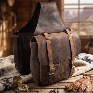 May include: Two brown leather saddlebags with buckles and straps. A compass, belt buckle, and leather gloves are on a wooden surface. The word "SAISHAS" is at the bottom of the image.