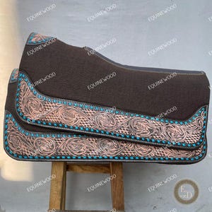 May include: A brown saddle pad with intricate copper-colored and turquoise decorative trim. The pad is displayed on a wooden stand, showcasing its detailed design and craftsmanship. The image is branded with the word "EQUINEWOOD".