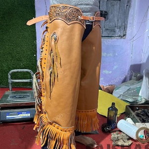 May include: Tan leather chaps with intricate floral tooling on the upper portion. The chaps feature decorative fringe along the sides and bottom, with additional embellishments. A belt with a buckle secures the chaps at the waist. The chaps are designed for riding.