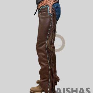 May include: Brown leather chaps featuring a floral-tooled leather yoke with turquoise piping. The side of the leg includes a row of turquoise studs and long leather fringe. The chaps are worn over blue denim jeans and tan leather boots.