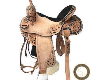 May include: A light tan leather western saddle with intricate floral tooling and a black seat. The saddle features silver-toned studs along the edges and is designed for horseback riding. The stirrups and straps are also visible.