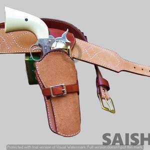 Peut inclure: Un revolver avec une poignée couleur crème et un cadre métallique est placé dans un étui en daim beige. L'étui comporte une sangle en cuir marron avec une boucle en métal. Une large ceinture en cuir beige avec des coutures blanches l'accompagne.