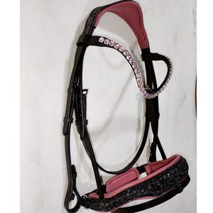 May include: A black leather horse bridle featuring a browband adorned with iridescent crystals. The headpiece and noseband are lined with soft pink padding, and the noseband is finished with a textured, black glittery surface for a distinct appearance.