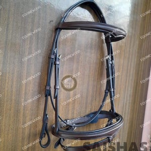 Puede incluir: Un cabezal de caballo de cuero marrón con una testera y una muserola acolchadas. El cabezal presenta correas ajustables, hebillas metálicas y un diseño curvo para la comodidad del caballo. La imagen está sobre un fondo de madera.