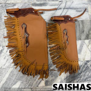 May include: A pair of tan leather chaps featuring dark brown floral-tooled leather yokes, long fringe along the sides, silver-toned concho accents, and adjustable buckle straps. The design includes rows of decorative stitching and metal hardware.