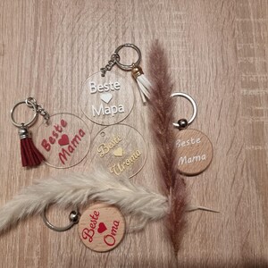 May include: A collection of keychains with the words "Beste Mama", "Beste Oma", and "Beste Uroma" in white and red lettering. Some keychains are clear acrylic, others are wooden. Each keychain has a metal ring and a tassel.