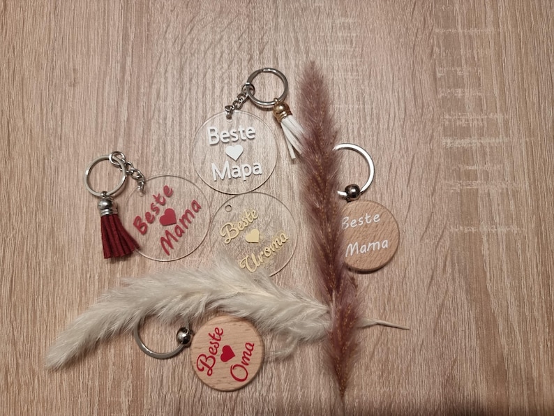 May include: A collection of keychains with the words "Beste Mama", "Beste Oma", and "Beste Uroma" in white and red lettering. Some keychains are clear acrylic, others are wooden. Each keychain has a metal ring and a tassel.