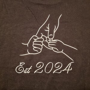 May include: Dark gray t-shirt featuring a white line drawing of three hands holding each other. The text "Est 2024" is written in a cursive font below the hands.