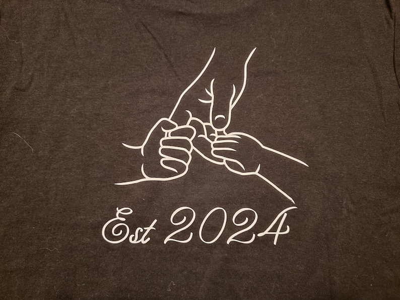 May include: Dark gray t-shirt featuring a white line drawing of three hands holding each other. The text "Est 2024" is written in a cursive font below the hands.