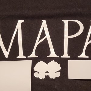 May include: Black t-shirt with the word "MAPA" in large white letters. Below the word are three white cutouts: a rectangle, a pair of connected cartoon figures, and another rectangle. The shirt is made of a dark fabric.
