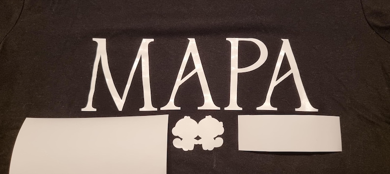 May include: Black t-shirt with the word "MAPA" in large white letters. Below the word are three white cutouts: a rectangle, a pair of connected cartoon figures, and another rectangle. The shirt is made of a dark fabric.