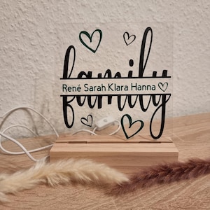 May include: A clear acrylic family sign with the word "family" in a cursive font, and the names "Ren&eacute; Sarah Klara Hanna" below. The sign is mounted on a wooden base with a power cord and a switch.