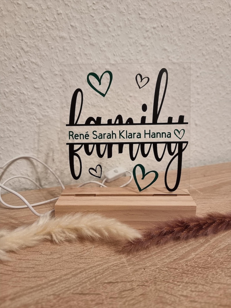 May include: A clear acrylic family sign with the word "family" in a cursive font, and the names "Ren&eacute; Sarah Klara Hanna" below. The sign is mounted on a wooden base with a power cord and a switch.