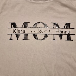 May include: White t-shirt with the word "MOM" in large, stylized black letters. Below the "O" is the name "Klara" on the left and "Hanna" on the right, separated by a heart symbol.