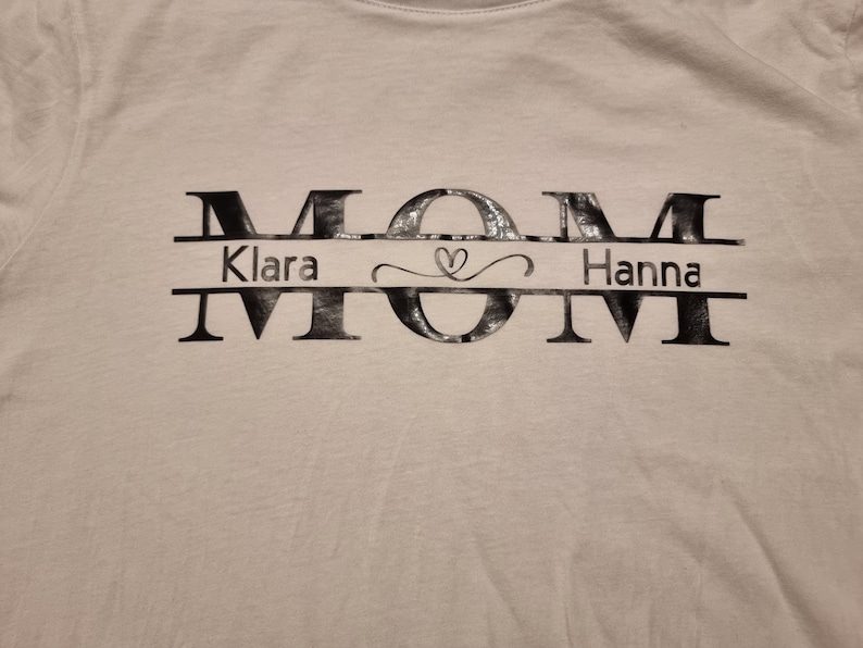 May include: White t-shirt with the word "MOM" in large, stylized black letters. Below the "O" is the name "Klara" on the left and "Hanna" on the right, separated by a heart symbol.