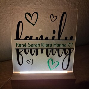 May include: A lighted acrylic sign with the word "family" in a script font, with the names Ren&eacute;, Sarah, Klara, and Hanna below. The sign has a wooden base and features heart-shaped accents in black and green.