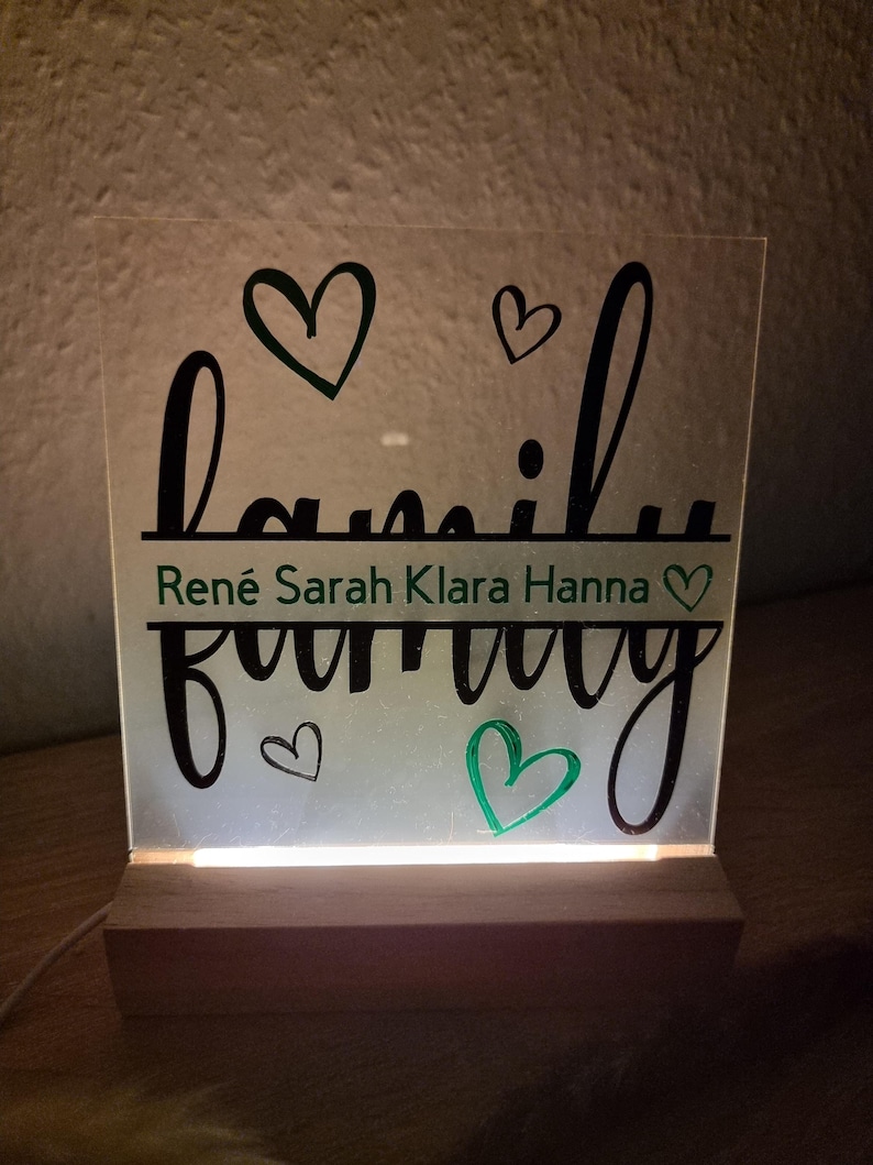 May include: A lighted acrylic sign with the word "family" in a script font, with the names Ren&eacute;, Sarah, Klara, and Hanna below. The sign has a wooden base and features heart-shaped accents in black and green.
