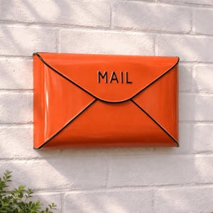May include: Orange metal mailbox shaped like an envelope, mounted on a white brick wall. The word "MAIL" is embossed on the front. Black trim outlines the envelope shape.