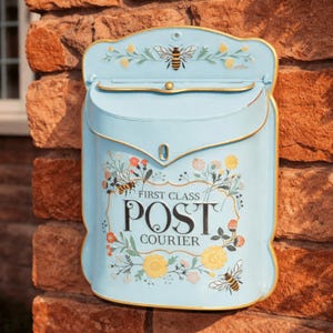 May include: A light blue metal mailbox with a decorative bee and floral design. The mailbox has the words "FIRST CLASS POST COURIER" printed on the front. The mailbox is mounted on a brick wall.